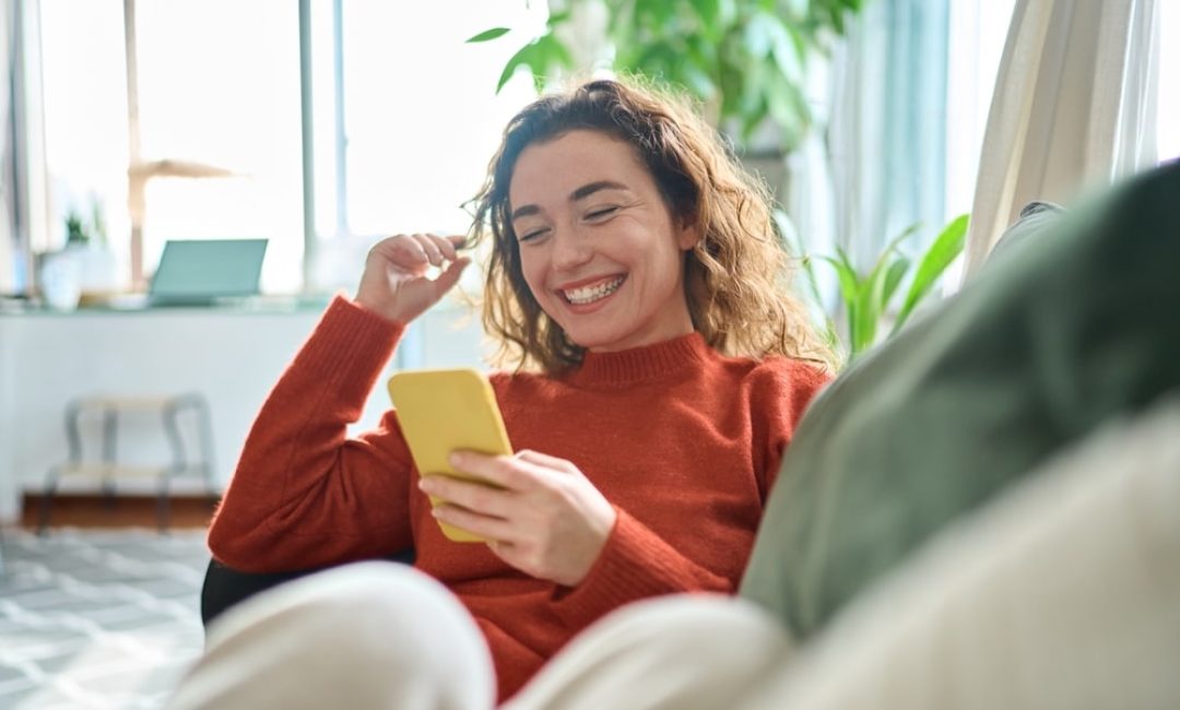 Happy,Relaxed,Young,Woman,Sitting,On,Couch,Using,Cell,Phone,